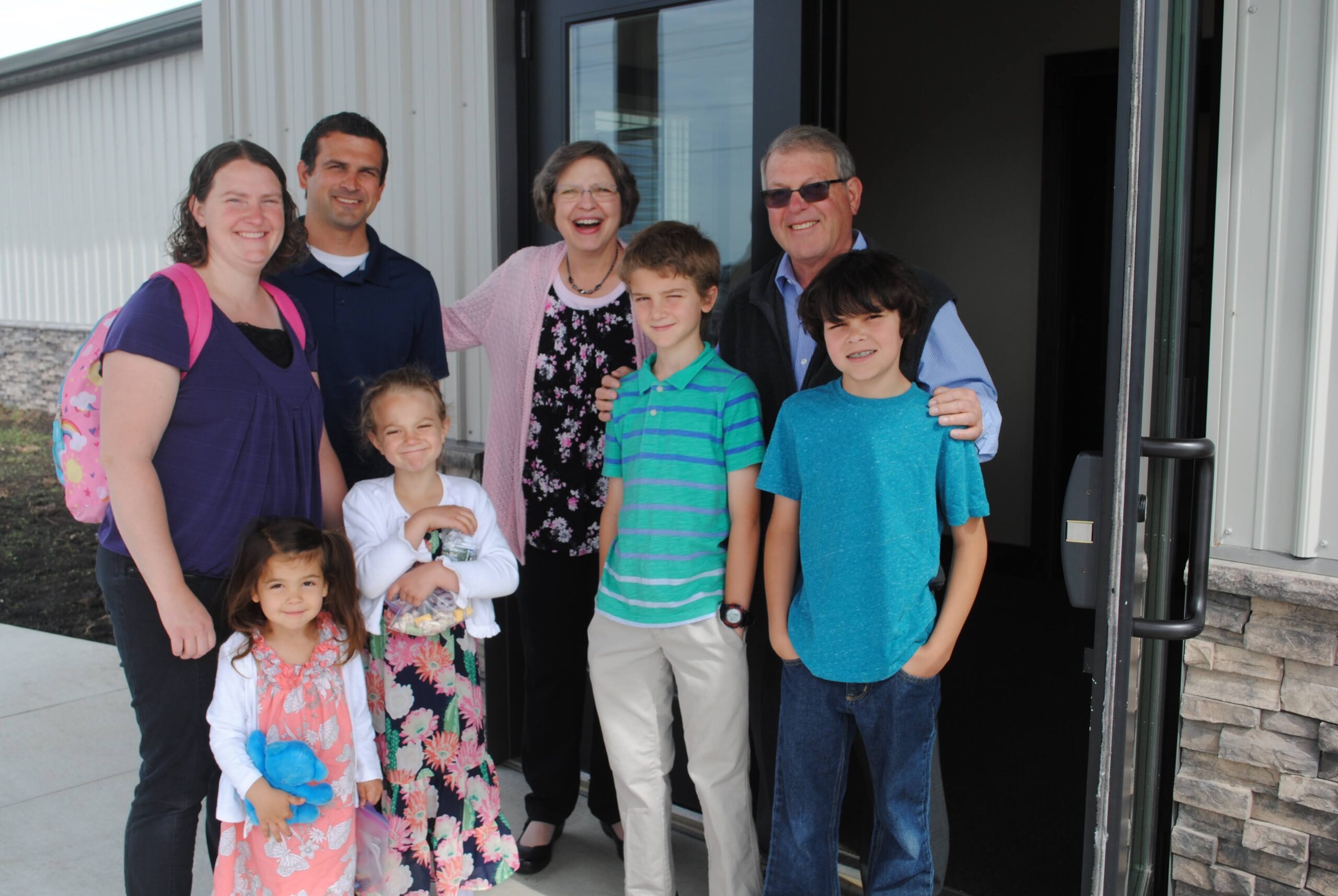 A family of grandparents, parents, and children gathered together outside the front door of the church smiling.